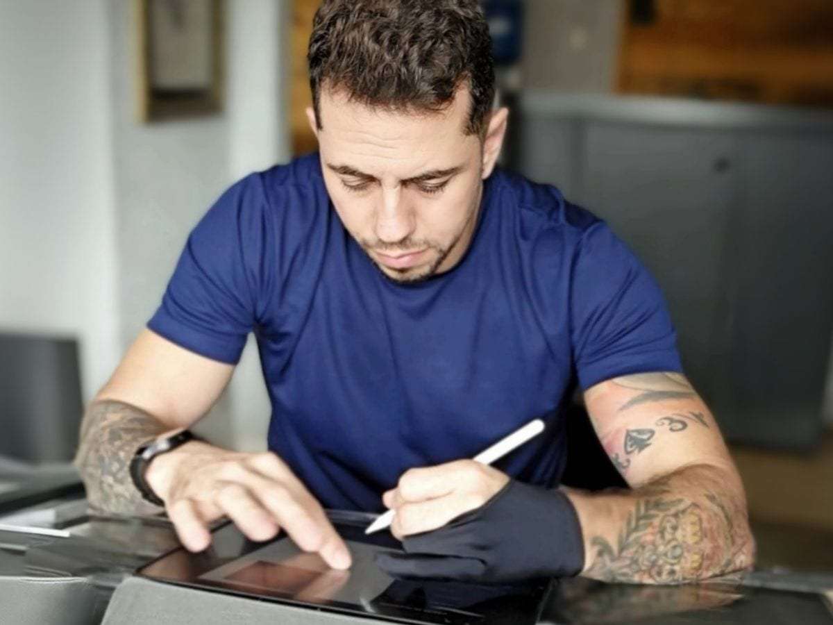Beto desenhando numa mesa digital, com os braços tatuados apoiados na mesa do estúdio