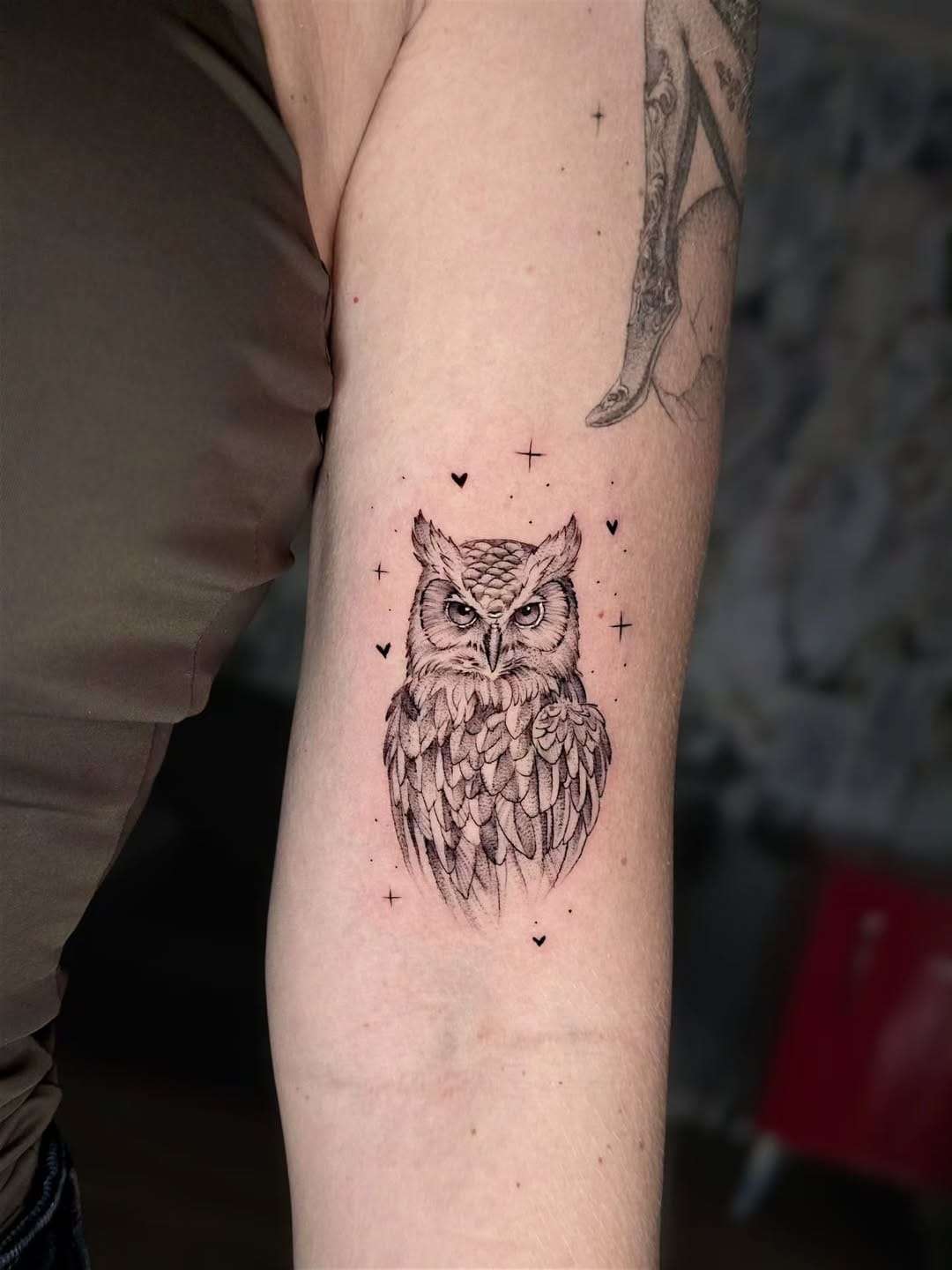 Tatuagem de uma coruja em fine line no braço, cercada de pequenas estrelas e corações